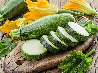 Eine Zucchini liegt auf einem Holzbrett. Daneben liegen mehrere Zucchini-Scheiben und Zucchini-Blüten.