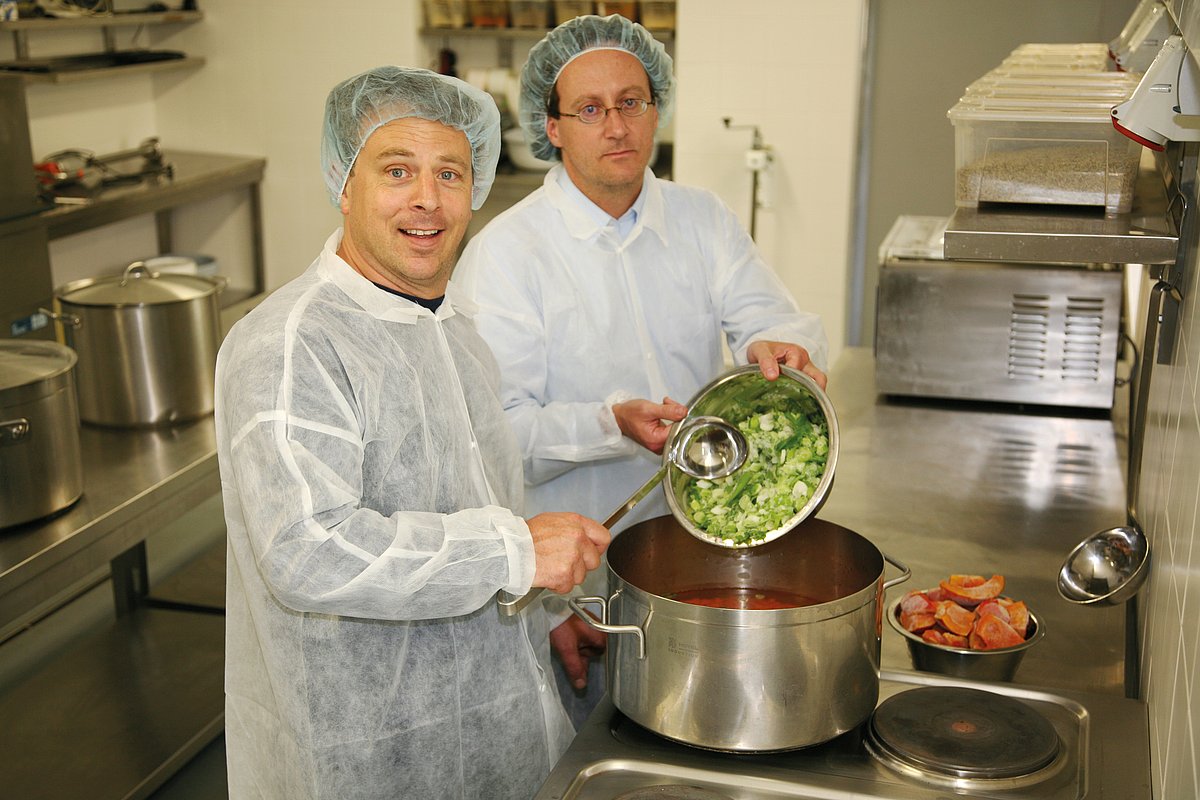 Zwei Köche in Arbeitskleidung bereiten in einem großen Topf Essen vor. 