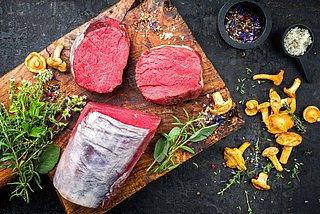 Ein Stück Rindfleisch liegt aufgeschnitten auf einem Holzbrett und ist garniert mit verschiedenen Kräutern und Gewürzen.