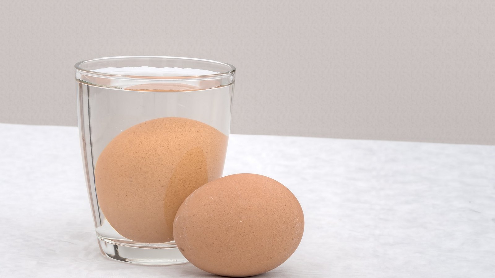 Ein braunes Ei liegt neben einem Glas, in dem ein weiteres braunes Ei in Wasser liegt.