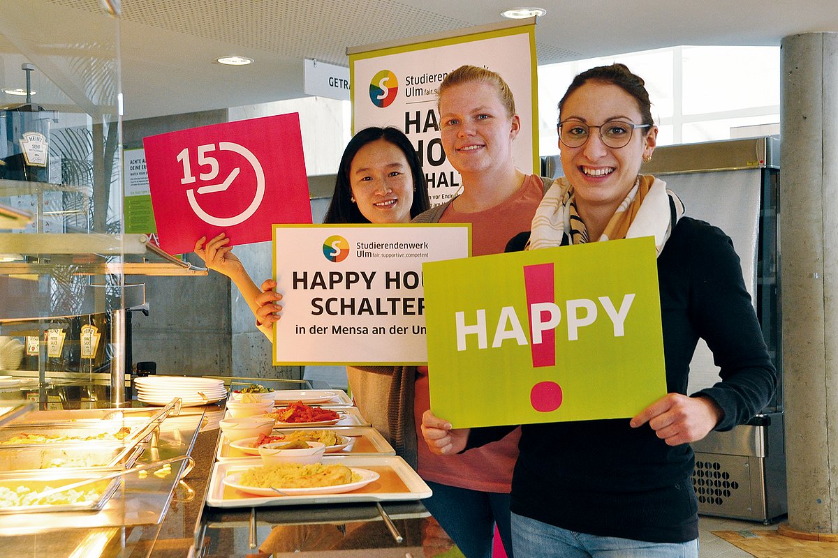 Drei Personen halten unterschiedliche Plakate in der Hand in einer Mensa. Auf einem Plakat steht "Happy Hour". 