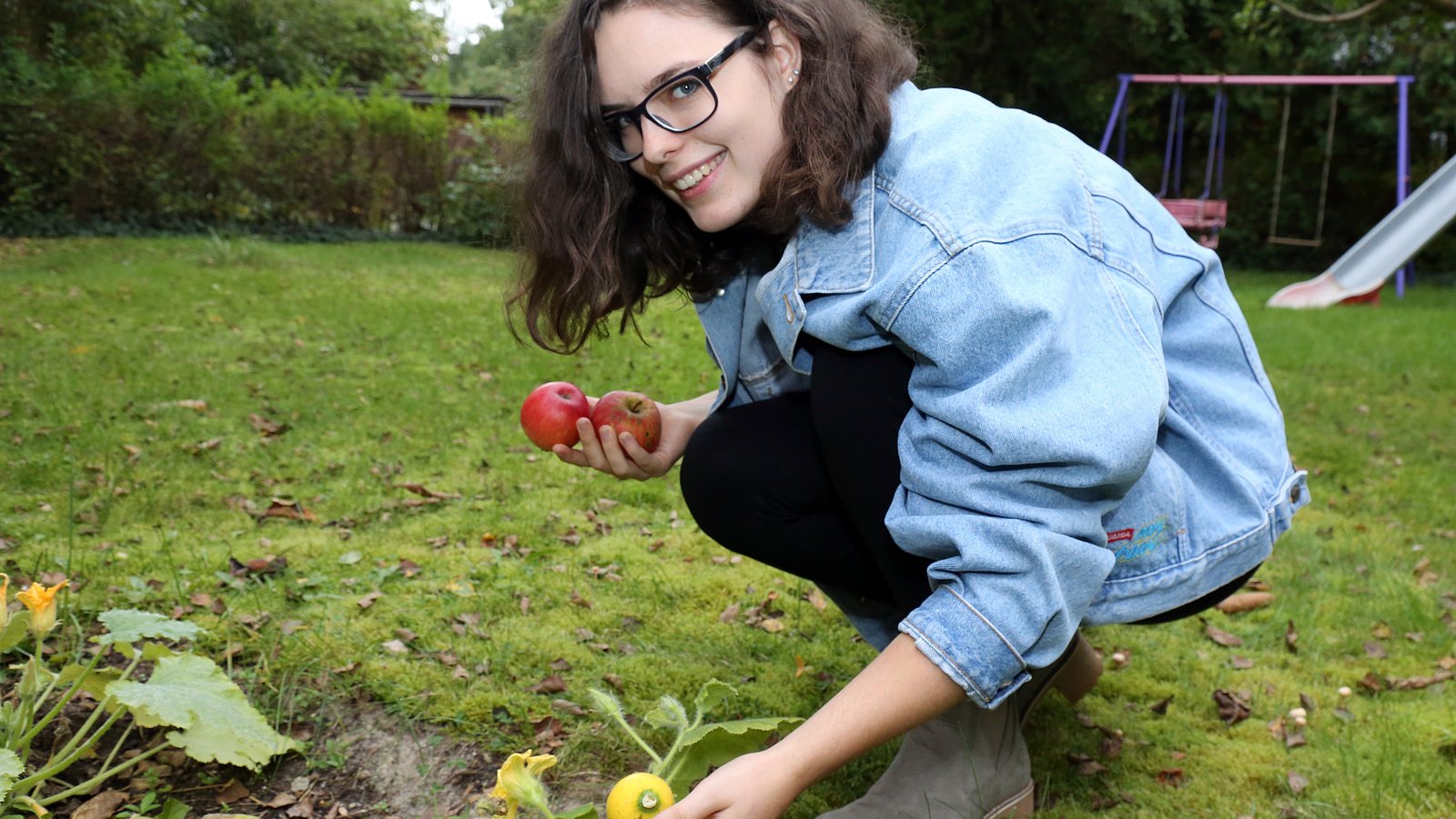 Die Gründerin hält zwei Äpfel in der Hand und kniet neben einem Gemüsebeet.