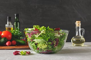 Salat wird in einer großen Glasschüssel präsentiert. Daneben stehen mehrere Flaschen mit Öl und Essig und weiteres Gemüse liegt auf einem Holzbrett.