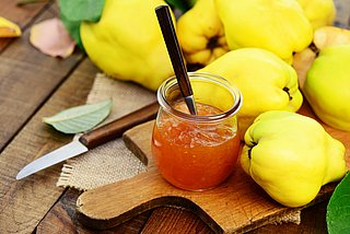 Gelbe Quitten liegen auf einem Holzbrett. Davor steht ein Glas mit Quitten-Marmelade und liegt ein Messer.