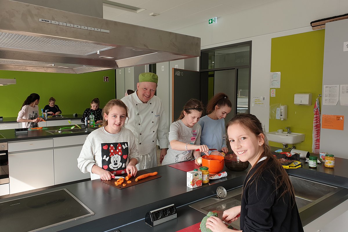 Kinder bereiten in einer Küche gemeinsam mit einem professionellen Koch Essen zu.