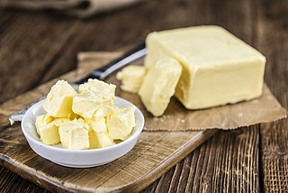 Ein Stück Butter liegt aufgeschnitten auf einem Holzbrett. Davor steht eine kleine Schüssel mit kleineren Butterstücken.