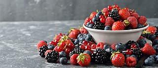 Eine Schale mit Brombeeren, Himbeeren, Erdbeeren, Blaubeeren und Johannisbeeren steht auf einem Tisch. Drum herum liegen ebenfalls Beeren.