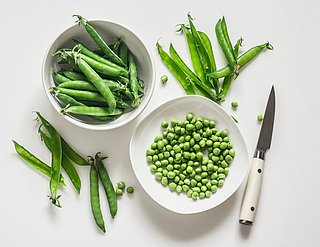 Eine Schüssel mit Erbsen und eine Schüssel mit Erbsenschoten. Daneben liegt ein Messer und weitere Erbsenschoten.