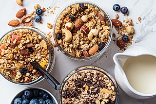 Verschiedene Müslisorten werden in drei Schüsseln präsentiert. Daneben steht eine Schüssel mit Brombeeren und ein Krug mit Milch.