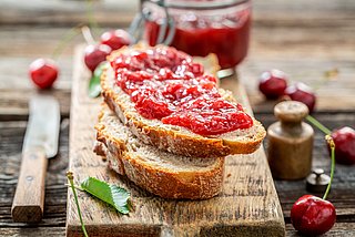 Zwei Brotscheiben bestrichen mit Marmelade liegen auf einem Holzbrett. Dahinter steht ein Einweck-Glas mit Marmelade und daneben liegt ein Messer.