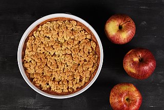 Ein Apple Crumble steht in einer Servierform auf dem Tisch, umgeben von drei Äpfeln