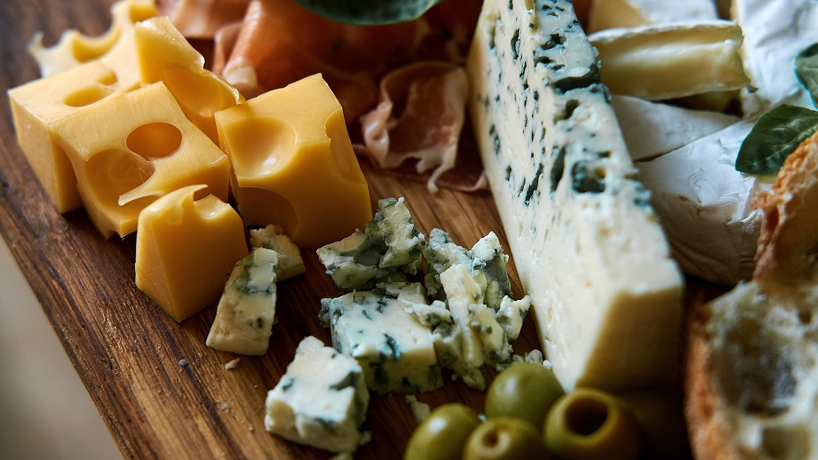 Verschiedene Käsesorten liegen auf einem Holzbrett neben Oliven.